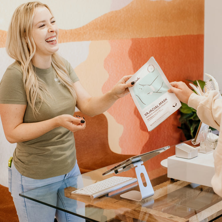 Woman smiling while handing a Vajacial Mask product brochure to a customer.